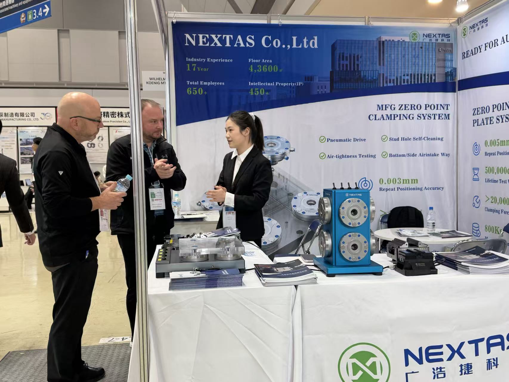 Tres personas conversando en la mesa de NEXTAS junto a un demo azul de varios puertos y placas redondas en cajas transparentes